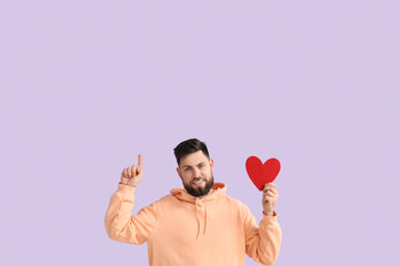 Young bearded man with paper heart for Valentine's Day pointing at something on lilac background