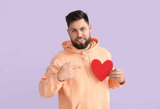 Young bearded man pointing at paper heart for Valentine's Day on lilac background