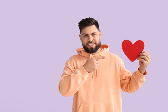 Young bearded man pointing at paper heart for Valentine's Day on lilac background - Powered by Adobe