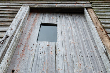 The exterior wall of an old weathered barn. The wooden walls and door are grey, textured, and worn. The door has vertical lines and the wall has horizontal boards. The door has a small glass window.