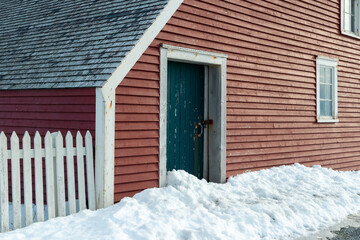 A closed small vintage green wooden shed door with a lock and latch. The barn has a vibrant red clapboard exterior wall with horizontal lines, white trim, and peeling paint with a mound of fresh snow. - Powered by Adobe