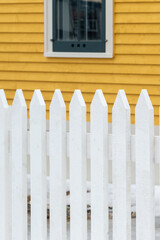 A yellow country style house with clapboard siding and a vintage double hung closed glass window with white trim behind a white picket fence. The exterior yellow wall is covered in rough boards. 