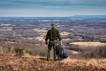 A hiker looking over a cliff from a hiking trail with a backpack, camping on a mountain bluff overlooking the valley with Winchester Tennessee in the background.