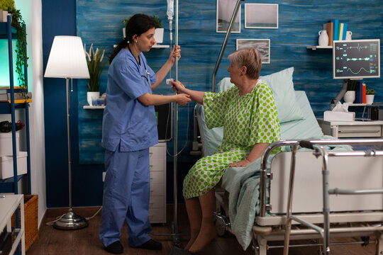 Female Skilled Nursing Home Nurse Assisting Elderly Lady In Room Receiving Intravenous Treatment. Senior Woman Patient With Chronic Illness Hospitalized In Health Care Facility