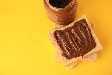 Tasty toast with chocolate paste and jar of cream on yellow background, above view. Space for text