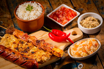 Espetinhos de churrasco em tábua de madeira com acompanhamentos, arroz, vinagrete, farofa e maionese