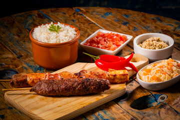 Espetinhos de churrasco em tábua de madeira com acompanhamentos, arroz, vinagrete, farofa e maionese