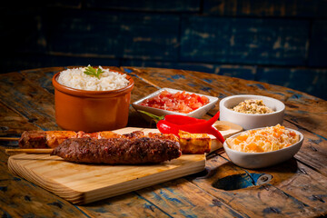 Espetinhos de churrasco em tábua de madeira com acompanhamentos, arroz, vinagrete, farofa e...