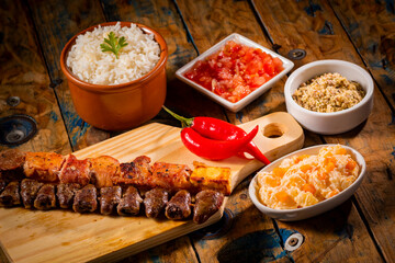 Espetinhos de churrasco em tábua de madeira com acompanhamentos, arroz, vinagrete, farofa e maionese