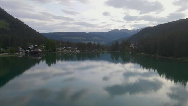 Aerial drone flight backwards over green lake Dobbiaco (Toblacher) in partly cloudy weather in calm waters.