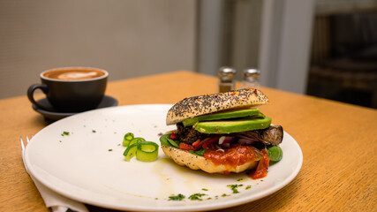 Vegan and vegetarian-friendly toasted chia seed sourdough bagel topped with avocado, shredded carrots, eggplant, red onions, sun-dried tomatoes, and spinach leaves with cappuccino on the side.
