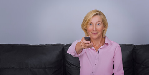 An attractive adult woman is sitting on the sofa with a remote control and switching channels on...