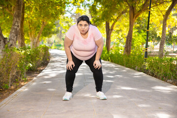 Overweight tired exhausted indian woman stooped after jogging workout outdoors in park. Fat lady relaxing after exercise. Fitness lifestyle. Full length Shot. - obrazy, fototapety, plakaty
