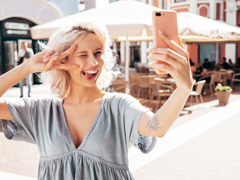 Young beautiful smiling hipster woman in trendy summer dress clothes. Sexy carefree model posing in the street at sunny day. Positive blond female outdoors. Cheerful and happy. Takes selfie photos