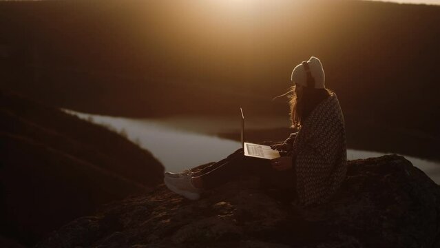A freelancer woman wrapped in a blanket working on the top of a mountain at sunset, after finishing her work closes her laptop and happily admires the nature landscape with an incredible panorama