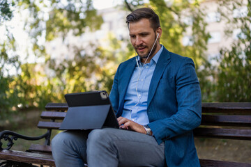 Caucasian male entrepreneur freelancer sitting outdoors in city park a bench talking online a video call using laptop or tablet. Business man office worker working on urban street background