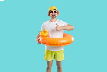 Vacation time. Cheerful guy with funny inflatable circle for swimming shows thumb up on light blue background. Joyful skinny young man in shorts, T-shirt and panama recommends summer vacation.