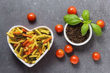 Raw pasta fusilli in heart bowl with cherry tomatoes and basil.