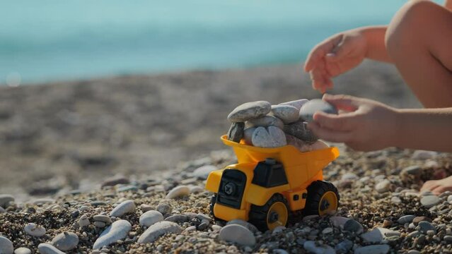 Close-up of a little boy's hands loading a toy truck with stones, standing on a sea beach. A small blond boy of 5 years old is playing on the beach near the sea with a toy truck. 4k footage