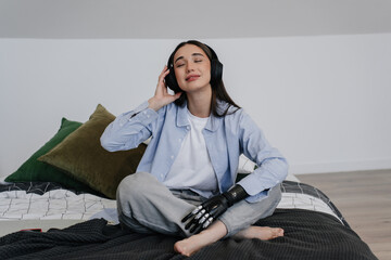 Young woman sitting on bed at home, with  bionic hand prosthesis, smiling, relaxing after remote...