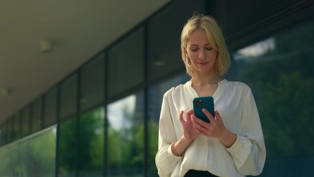 Attractive young business woman wearing formal suit standing near office centre background using the mobile phone, texting with clients, swiping apps. People and technology concept.