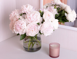 de-focused frame with a bouquet of light peonies in a vase and a candle on the dressing table. Blurred reflection of the bouquet in the mirror