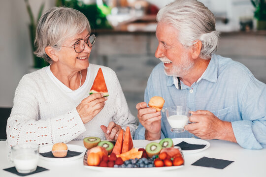 Cheerful beautiful senior couple at home having breakfast together with muffin, milk and fresh seasonal fruit, healthy eating concept