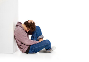 Portrait of young boy sitting on floor in depression and sadness over white background. Problems of youth