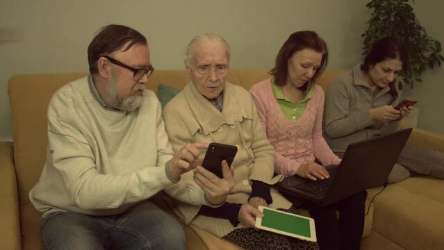 Family members use smartphones, tablet pc ( green screen ) and laptop. Slow motion 2x. An elderly man, a grandmother, a middle-aged woman and a young woman are sitting on a sofa in a room. 