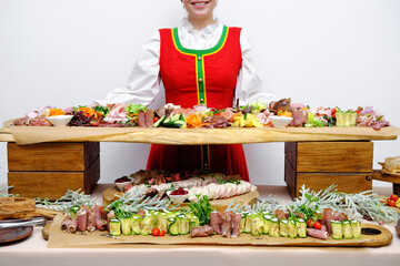 Beautiful smiling russian girl in folk costume stands near the buffet table and offers snacks