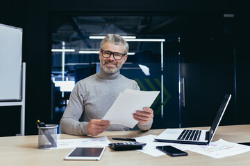 Successful businessman with documents smiling and looking at camera, portrait of mature gray-haired investor financier inside modern office, man behind paper work with reports accounts and contracts
