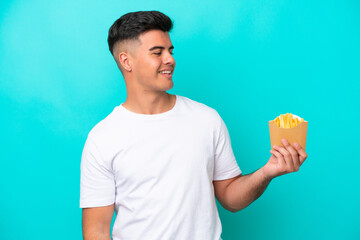 Young caucasian man catching french fries isolated on blue background looking to the side and smiling