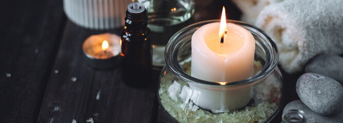 Spa setting with essential oil, candle, sea salt, pebbles, towel on dark wooden background. Massage, aromatherapy. Natural organic ingredients for relaxation, detention, wellness. Banner copy space