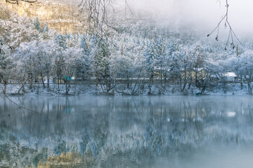 Karst deep-water lake in the mountains. Lake in winter with a view of snow-covered trees. Trees in the snow growing on the mountainside. Clean water in a non-freezing mountain lake. Fog on the lake.