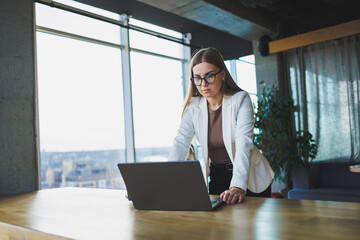 Smiling young woman in casual clothes and glasses, wearing a white jacket and laughing while sitting at a table with a laptop while working. Work in the office