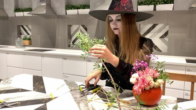 A girl in a witch costume and with a painted face creates a flower composition from a pumpkin. High quality photo