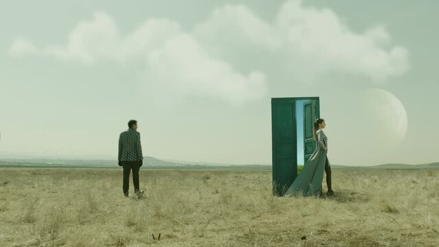 Couple walking on field near door . Young stylish man and woman stand on meadow field . Futuristic Fashion concept . Surrealistic scene . Way to another planet . Planets at day in the sky . Surrealism