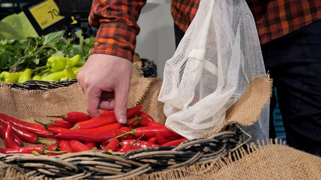 Male salesman fills red chili pepper into reusable bag. Sale of eco products at the farmers' market, organic vegetables at the local farmers' fair. Buying hot chili peppers in the vegetable section