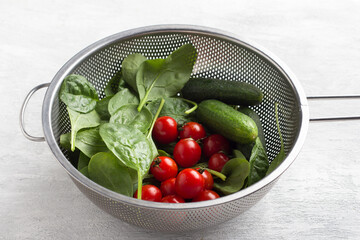 Fresh washed spinach leaves, cherry tomatoes and cucumbers in a colander on a light gray background. Preparing healthy homemade salad - 558913619