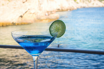 A blue cocktail drink in a tall glass against a blurred background of blue water at the beach