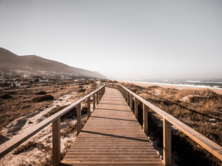 Obraz premium A long boardwalk on a grassy sand dune along the beach leading to the mountain with desaturated colors. The way ahead concept. Quiaios Beach, Portugal