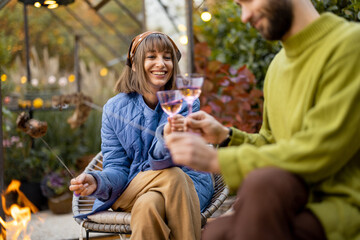 Young stylish couple grilling food and warming up while sitting together by the fire, spending autumn evening time at cozy atmosphere in garden