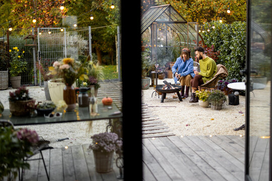 Young stylish couple grilling food and warming up while sitting together by the fire, spending autumn evening time at cozy atmosphere in garden. Wide view through the window