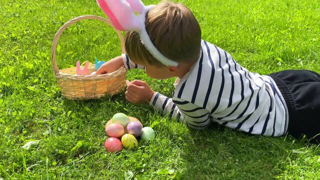 A boy with hare ears lies on the grass with colorful eggs. Egg hunting is an Easter tradition.
