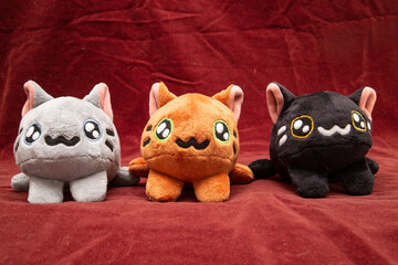 Three multi-colored plush cats are hand-sewn in the form of a soft toy and lie facing the camera against a dark red background.