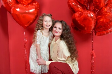 beautiful little girl hug with mom, mother smiles on red background with heart-shaped balloon and a gift box. The concept of love, present, Valentine's Day, February 14, March 8, mother's day