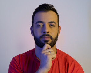 Homem moreno de cabelo curto e barba vestindo uma camiseta laranja com a mão no queixo