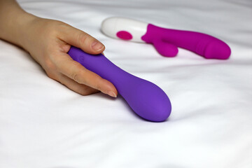 Sex toys in female hand on white sheet. Woman lying on a bed with purple silicone dildo and red vibrator - obrazy, fototapety, plakaty