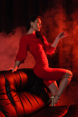 side view of brunette woman in red dress posing near couch on black with smoke.