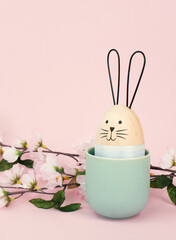 Cute easter bunny or rabbit with a smiling egg face in a cup, spring holiday greeting card with cherry blossoms

